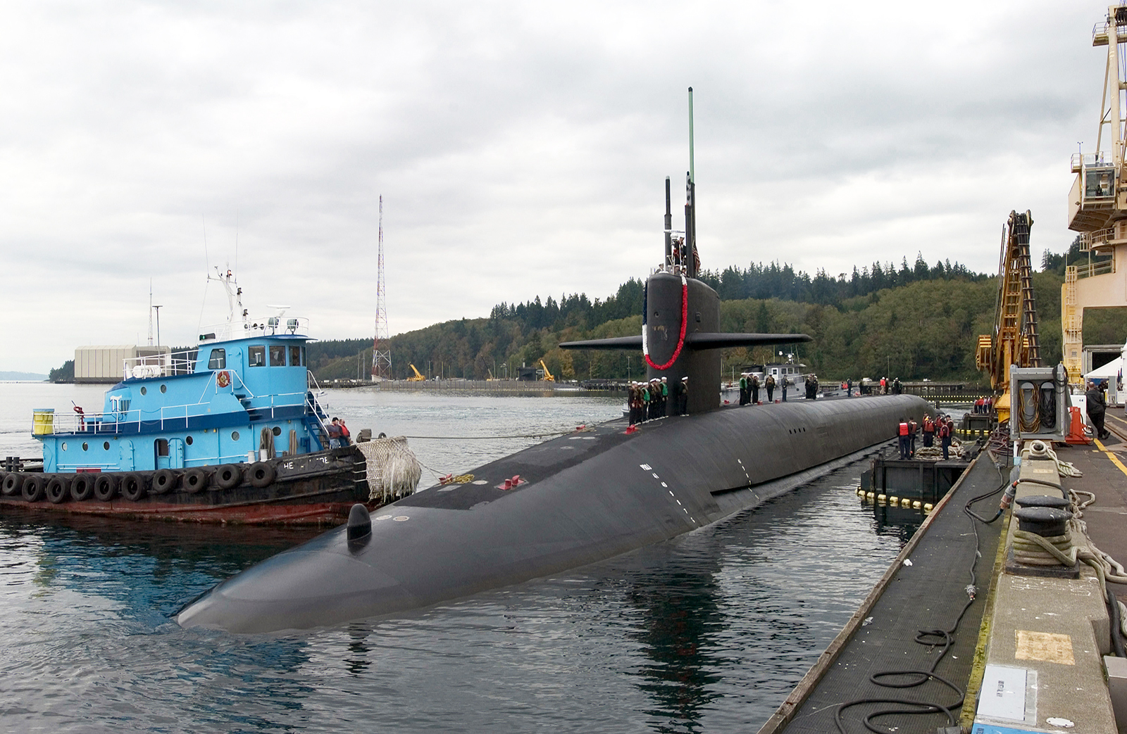 A starboard bow view of the US Navy (USN) OHIO CLASS: Strategic Missile Submarine, USS NEBRASKA (SSBN 739), showing Sailors manning the rails as the ship is assisted by a commercial tugboat, while mooring at its new homeport for the first time at the Delta Pier located at Naval Base Kitsap–Bangor, Washington (WA).  (Duplicate image, see also DNSD0610169 or search 041020N6497N044)