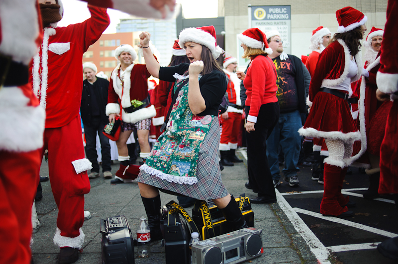 A Mrs. Claus stands over a set of boomboxes while dancing.