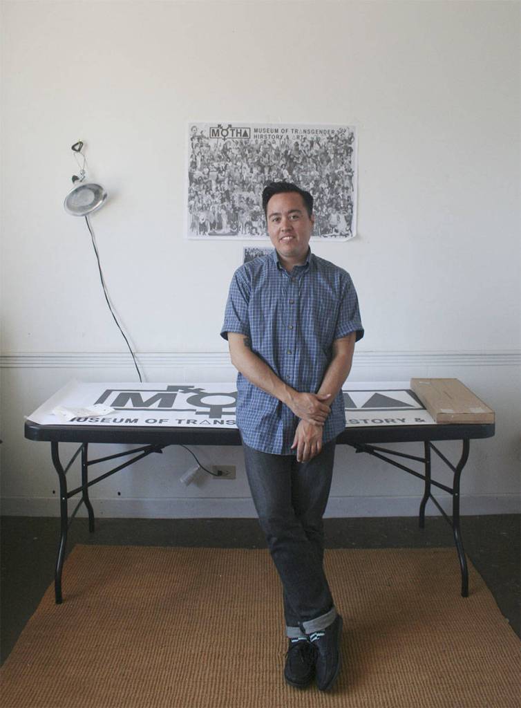 Chris E. Vargas stands in front of a Museum of Trans Hirstory & Art poster and banner. Photo by Michele Carlson