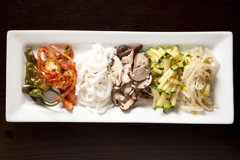 Assigning a charitable fee to banchan flips the meaning of the snacks, which are typically given freely and generously as a signal of hospitality. It's not unusual for a Korean restaurant to present its guests with six to eight complimentary rounded stainless-steel bowls brimming with tangles of spicy mung bean sprouts, shredded dried squid, cubes of snow-white pickled radish, and braised potatoes. When a bowl's emptied, it's returned to the kitchen for a refill. Chan, by contrast, serves a rectangular white plate set with two dainty chopstick scoops apiece of whole shishito peppers, marinated mushrooms, carrots, zucchini and sprouts. No seconds.