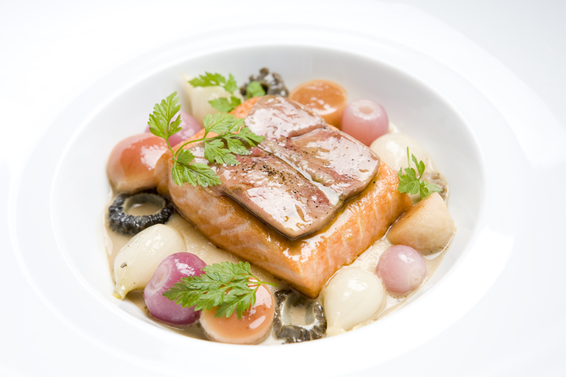A hunk of ivory-fleshed salmon crowned with a slice of foie gras is sunk in a shallow bowl, tightly surrounded by pearl onions and mushrooms.