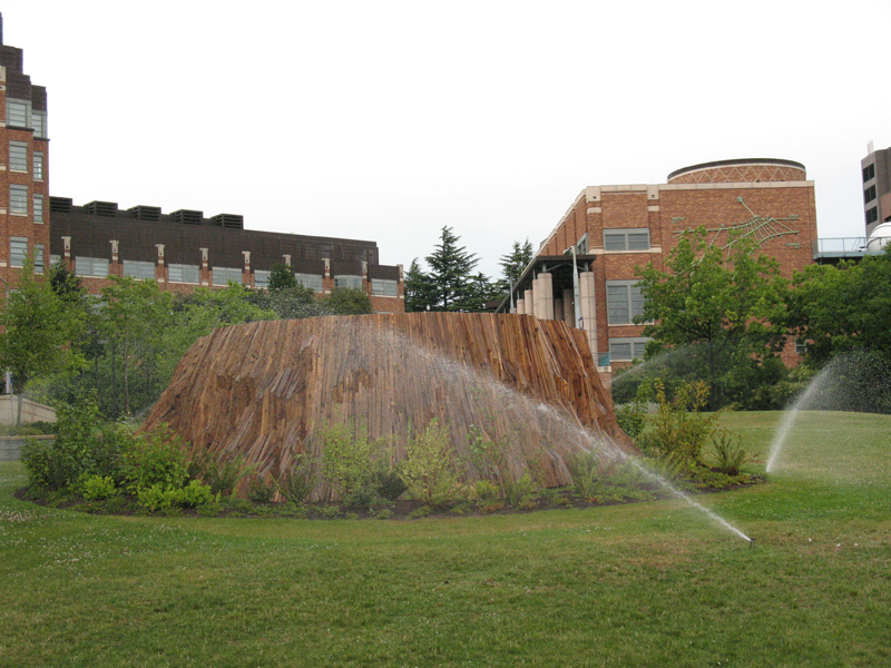 Stronghold, UW campus, NE Pacific St. at 15th Ave. NE.: The original UW campus downtown was once covered with old-growth timber, as was its present location when the school moved north in 1895. Now, as that institution continues its inexorable sprawl south of Pacific Avenue toward Portage Bay, New York artist Brian Tolle reminds us of that arboreal past with his recently installed Stronghold. It is, at first glance, nothing more than stump on a manicured lawn. Walk closer (sprinklers permitting), and youaE™ll see that itaE™s a constructed stump, an invented artifact made of inexpensive cedar slats. Measuring about 23 feet in diameteraE”with a seating area within, bring your lunchaE”Stronghold suggests the enormous tree trunks that once drove this regionaE™s economy, that helped establish the city and our stateaE™s first university (founded in 1861). But such towering cedars and firs are all gone, of course, and TolleaE™s materials are of the size and grade you could buy at any lumber yard. This neo-stump stands next to a new UW bioengineering building onaE”appropriate, since technology is the new timber of the Northwest. In shape, the irregular ring also echoes our skyline of ancient, crumbling volcanoes (Mount St. Helens in particular) that were formed in violence. The installation recreates our local history before it met the crosscut saw.