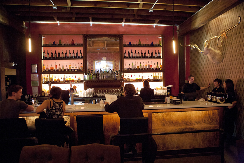 The vintage back bar and a server sideboard were salvaged from a drug store, but the room's most striking elements are brand new, including the slick white tables and bar top, and the wallpaper, red as papal slippers, imprinted with a gold pattern based on the gun-toting white ram.