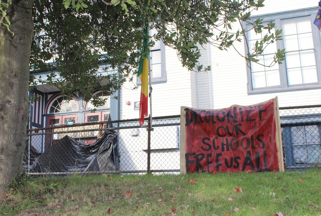 A chain-link fence decorated in blood-red banners bearing messages like DECOLONIZE OUR