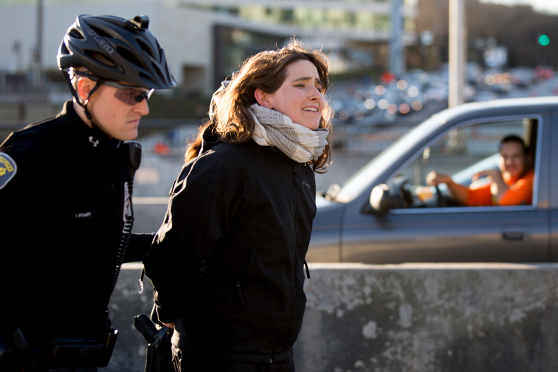 Freed from her device, a protester is led away by police while a driver passing by in the southbound lanes slows to watch.