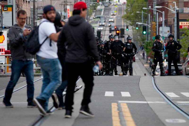Crowds survey the scene while crossing the street as cops in riot gear look on.